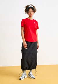 Tricou roșu din bumbac cu mâneci scurte și logo alb, asortat cu o fustă lungă neagră cu șnur, șosete gri și adidași cu model.
