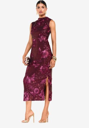 Mujer con vestido sin mangas de lentejuelas burdeos con estampado floral y abertura lateral, sosteniendo un pequeño bolso de mano y llevando tacones altos dorados y pulseras.