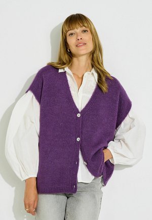 Mujer con cabello rubio largo lleva un cárdigan morado sin mangas con botones sobre una blusa blanca con mangas abullonadas y pantalones gris claro.
