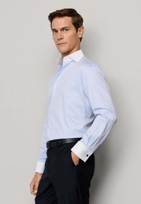 Mężczyzna o krótkich brązowych włosach, ubrany w jasnoniebieską koszulę z białym kołnierzykiem i mankietami, czarny pasek i ciemne spodnie, stojący na tle gładkiej ściany.