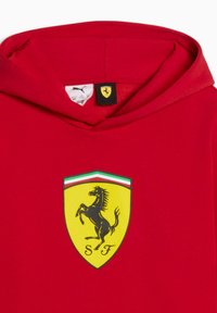 Sweatshirt à capuche rouge avec un logo Ferrari noir sur un écusson jaune, souligné par des bandes vertes et blanches en haut. Texture de tissu doux.