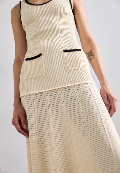 Top sin mangas de canalé color crema con ribete negro y dos bolsillos frontales, combinado con una falda de longitud a la rodilla con un patrón perforado.