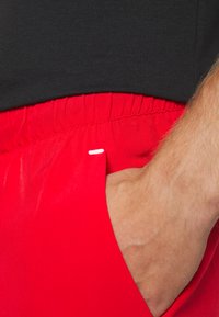 Červené šortky s elastickým pásom, majú bočnú kapsu a malý detail s bielou nášivkou. Hladká, ľahká tkanina.