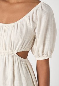 Witte structuur jurk met een vierkante halslijn en pofmouwen. Kenmerken zijn uitsnijdetails bij de taille en geplooide elastische accenten.