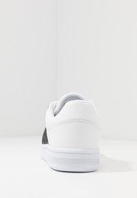 White sneaker with a smooth leather finish and textured black mesh side panel. Rubber sole with horizontal ridges, rounded heel.