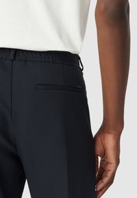 Marineblauwe broek met elastische tailleband, zijzak, gestructureerde stof en een klein metalen logo-detail op de achterkant. Regular fit ontwerp.