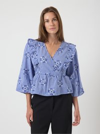 Blouse en vichy bleu avec des épaules volantées, broderie florale blanche et bleue, décolleté en V, taille peplum et manches longues en forme de cloche.