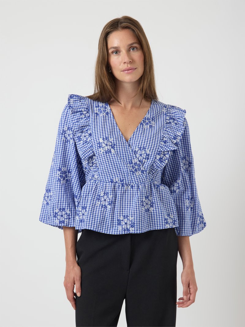 Blouse en vichy bleu avec des épaules volantées, broderie florale blanche et bleue, décolleté en V, taille peplum et manches longues en forme de cloche.