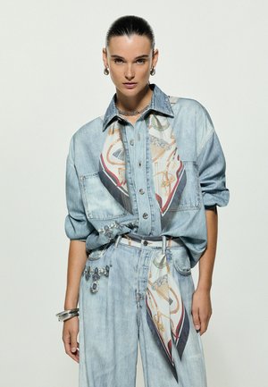 Femme portant une chemise et un pantalon en denim bleu clair avec des détails imprimés foulard, un collier chaîne argenté, des boucles d’oreilles créoles et des bracelets argentés, posant devant un fond blanc.