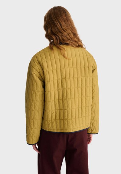 Personne aux longs cheveux ondulés portant une veste matelassée jaune moutarde et un pantalon foncé, vue de dos sur un fond clair uni.