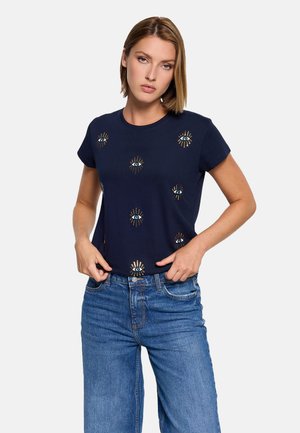 Mujer con camiseta azul marino con diseños de ojos y pantalones vaqueros azules, de pie con las manos cerca de la cintura contra un fondo liso.