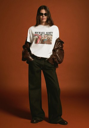 Nő napszemüvegben, fehér ingben "Michael Scott" felirattal és grafikával, barna bőrkabáttal a vállán, széles fekete nadrágban.
