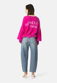 Rozā pūkaina džemperis ar lielu "RUMELI STUDIO" tekstu uz muguras, apvienots ar gaiši ziliem augsto jostasvietu džinsiem un caurspīdīgām papēža čībām.
