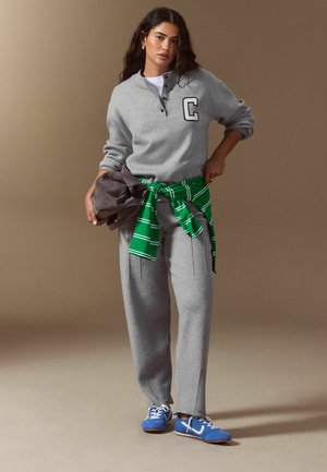 Frau trägt einen grauen Pullover mit dem Buchstaben C, graue Hose, blaue Sneakers und ein grün gestreiftes Hemd um die Taille gebunden, hält eine braune Jacke.