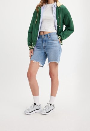 Kvinde iført grøn jakke over lagdelte skjorter, hvid cropped top, denimshorts med frynset kant, hvide sokker og hvide sneakers.