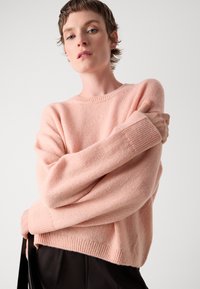 Personne à cheveux courts portant un pull en laine rose clair et un pantalon noir, tenant un sac noir contre un fond gris uni.