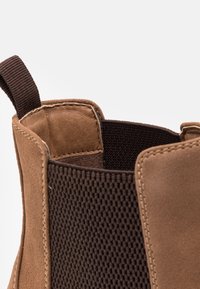 Gros plan sur une botte courte en daim marron avec panneau latéral élastique et languette pour un enfilage facile.