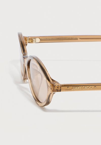 Διαφανή μπεζ στρογγυλά πλαίσια γυαλιών με καθαρούς φακούς και χρυσούς βραχίονες με χαραγμένο το λογότυπο "JIMMY CHOO" σε άσπρο φόντο.