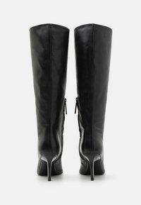 Knæhøye svarte lærstøvler med en glatt tekstur, spisse tåpartier og tynne høye hæler. Har to glidelåser på siden for lukking.