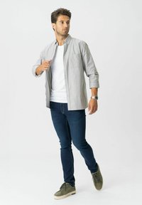 Férfi fehér pólót, világosszürke inget, sötétkék skinny farmert és oliva zöld sportcipőt visel, oldalra nézve sétál.