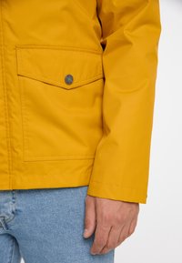 Giacca impermeabile gialla con vestibilità rilassata, caratterizzata da una grande tasca frontale e dettagli con bottoni a pressione sulla tasca. Indossata sopra jeans in denim blu chiaro.