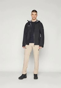 Svart lett zip-up jakke, beige cargobukser, marineblå t-skjorte og svarte sko. Jakken har sidelommer med glidelås og logo på brystet.