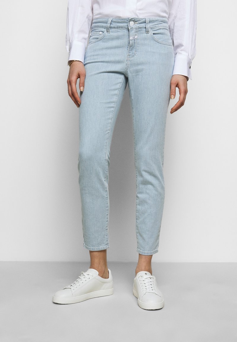 Light blue striped jeans with a slim fit, five-pocket design, and a button fly, paired with white sneakers. Fabric has a smooth texture.