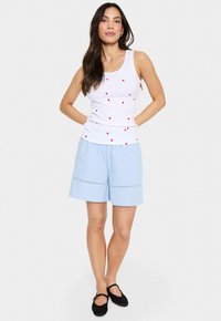 Top blanco sin mangas con patrones de corazones rojos dispersos, combinado con pantalones cortos azul claro con una costura decorativa. La modelo lleva zapatos planos negros.