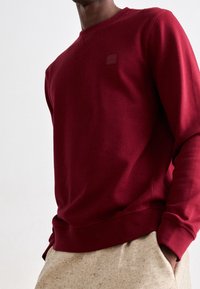 Sweatshirt bordeaux à manches longues, poignets côtelés et col rond ; présente un petit logo texturé sur la poitrine. Texture de tissu douce.