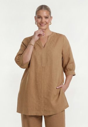 Frau mit blondem Haar in einer hellbraunen langärmeligen Tunika und passenden Hosen, lächelnd, mit einer Hand nahe am Kinn und der anderen in der Tasche.