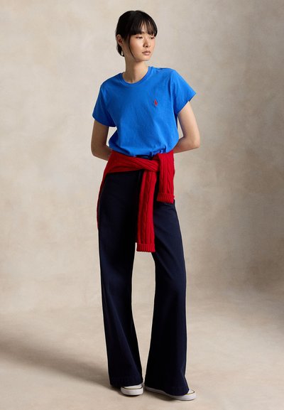 Junge Person steht, trägt ein blaues T-Shirt, roten Pullover um die Taille gebunden, schwarze weite Hosen und weiße Sneakers, schaut seitlich.