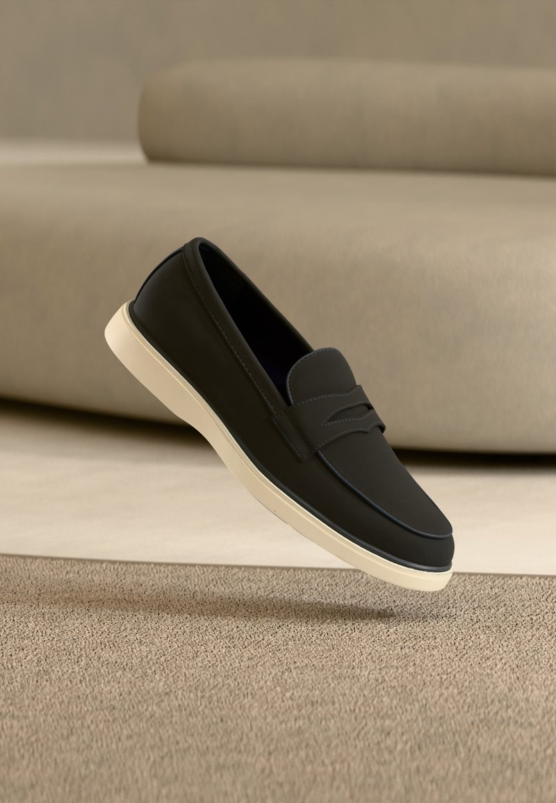 Mocassin noir à enfiler avec semelle blanche posé sur un tapis beige devant un canapé assorti.