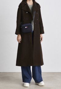 Lång brun kappa, blandning av ull, dubbelknäppt design, stor krage, kombinerad med en grön tröja, blå vida jeans och vita sneakers.