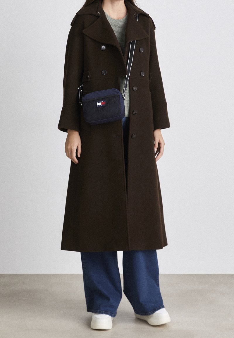 Lång brun kappa, blandning av ull, dubbelknäppt design, stor krage, kombinerad med en grön tröja, blå vida jeans och vita sneakers.