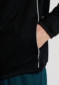 Mano appoggiata nella tasca di un giubbotto nero con piping bianco e un piccolo etichetta multicolore, indossando pantaloni turchesi.