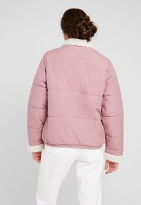 Lyserød puffjakke med en shearlingkrave, lodret quilted tekstur og en cropped design. Har lange ærmer og elastiske manchetter.