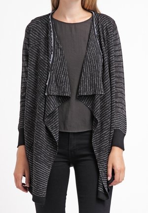 Femme portant un cardigan ouvert à rayures noires et grises sur un t-shirt gris foncé uni et un pantalon noir, les mains détendues sur les côtés.