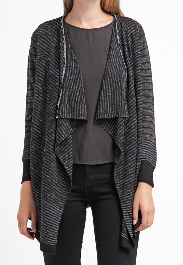 Femme portant un cardigan ouvert à rayures noires et grises sur un t-shirt gris foncé uni et un pantalon noir, les mains détendues sur les côtés.