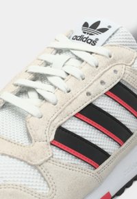 Detailný záber na bielu tenisku Adidas s béžovými semišovými detailmi, bielymi šnúrkami, čiernymi a červenými bočnými pruhmi a logom Adidas na jazyku.