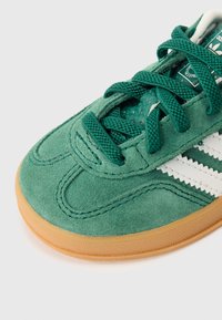 Žalios spalvos zomšiniai sportbačiai su tekstūruotu viršumi, baltomis juostelėmis ir gumine pado dalimi. Turi storas raištelius ir paminkštintą apykaklę.