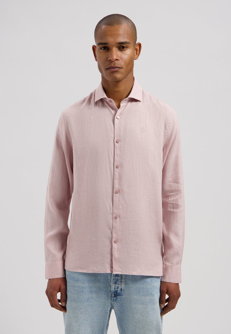 Homme portant une chemise à manches longues rose clair boutonnée et un jean bleu clair, debout devant un fond gris uni.