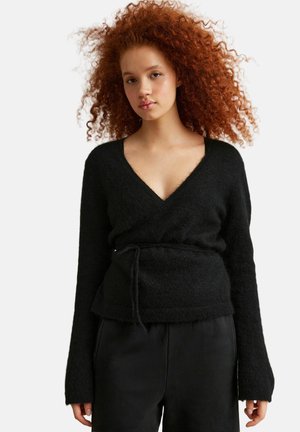 Młoda kobieta o bujnych, kręconych rudych włosach, ubrana w czarny sweter typu wrap i czarne spodnie na jednolitym tle.