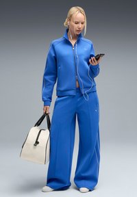 Chándal azul que incluye una chaqueta con cremallera y pantalones de pierna ancha, hecho de tela suave con ribetes blancos. Lleva un bolso crema con detalles en negro.