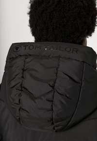 Veste rembourrée noire avec un col haut, des coutures visibles et un logo "TOM TAILOR" brillant dans le dos. Texture douce et matelassée.