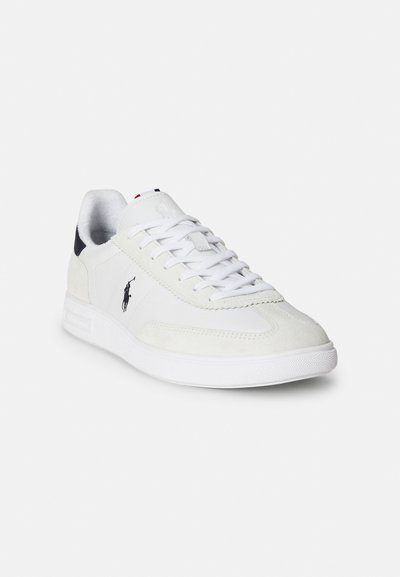 Vit sneaker med ovandel i mocka och läder, texturerad tåhätta, platta skosnören och en logotyp på sidan. Gummisula med mönster för grepp.