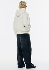 Überdimensioniertes cremefarbenes Kapuzenpullover mit Kordelzug, kombiniert mit weit geschnittenen, dunklen Jeans aus Denim mit Gesäßtaschen und einem leicht fransigen Saum.