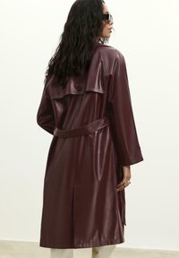 Burgundská eko kožená trench coat s opaskom v páse, širokými rukávmi a gombíkovou zadnou časťou. Hladká textúra s minimálnym zdobením.