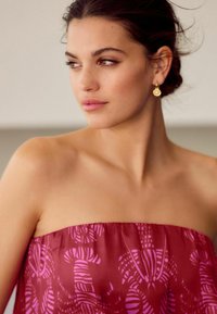 Strapless jurk in diep bordeaux met een roze abstracte patroon, zachte stof en elegante val. Gouden oorbellen voegen een subtiele accenten toe.