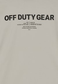 Beżowa bawełniana koszulka z odważnym czarnym napisem "OFF DUTY GEAR" oraz różnymi nadrukowanymi detalami i symbolami. Miękka faktura, standardowy krój.