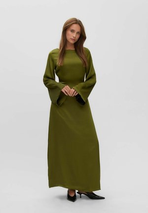 Femme aux longs cheveux bruns portant une longue robe vert olive à larges manches et des talons noirs pointus, debout devant un fond uni.